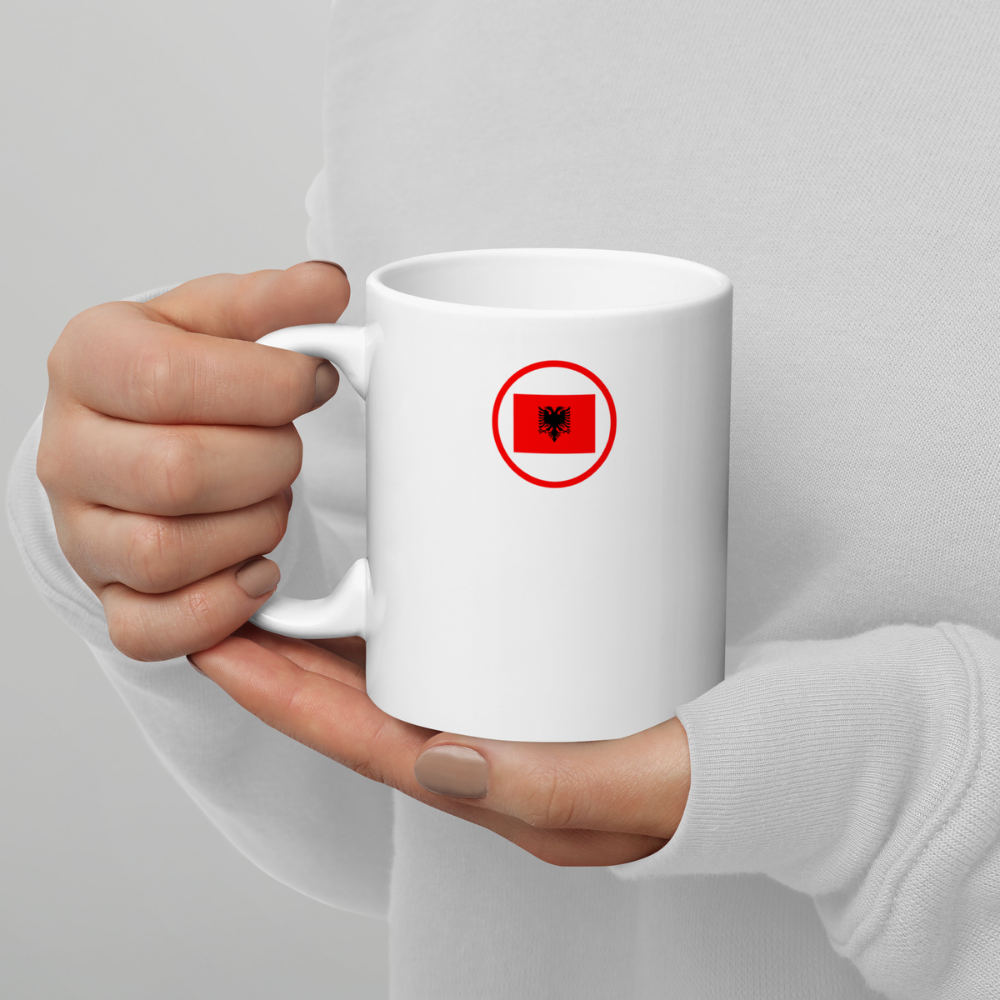 Mug blanc tenu à deux mains avec emblème albanais Personne tenant un mug en céramique blanche avec les deux mains, présentant un petit logo circulaire rouge contenant l’aigle noir à deux têtes du drapeau de l’Albanie.