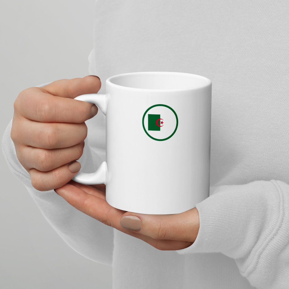 Mug blanc tenu en mains avec emblème du drapeau algérien Personne tenant un mug en céramique blanche avec un emblème circulaire du drapeau algérien imprimé sur la face avant.